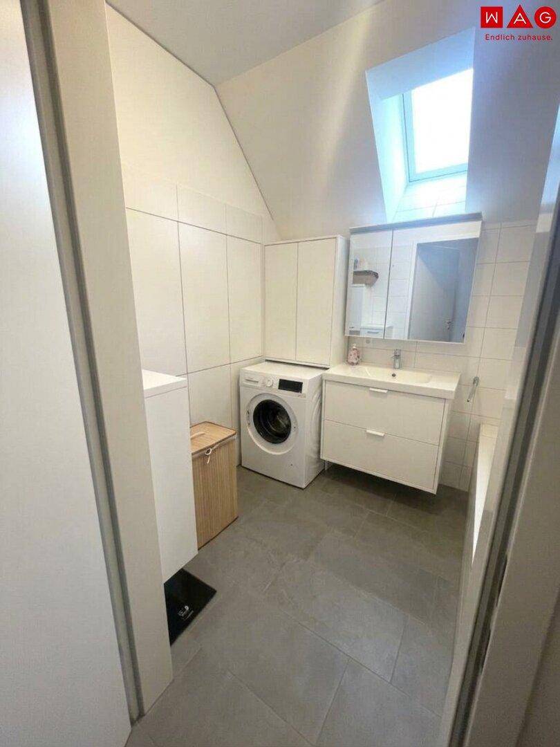 Modernes Badezimmer mit Waschmaschine, Waschtisch und Spiegelschrank, beleuchtet durch ein Dachfenster.