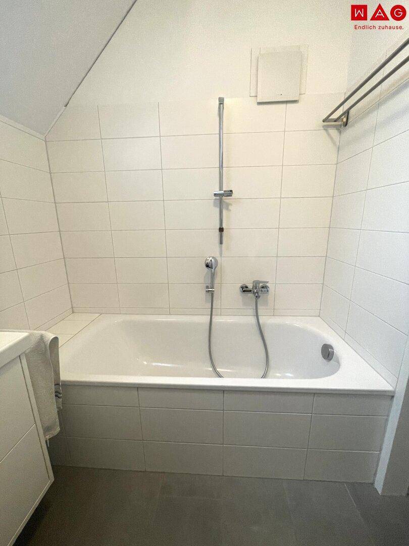 Eingebaute Badewanne mit Duscharmatur in einem hell gefliesten Badezimmer mit schräger Decke.