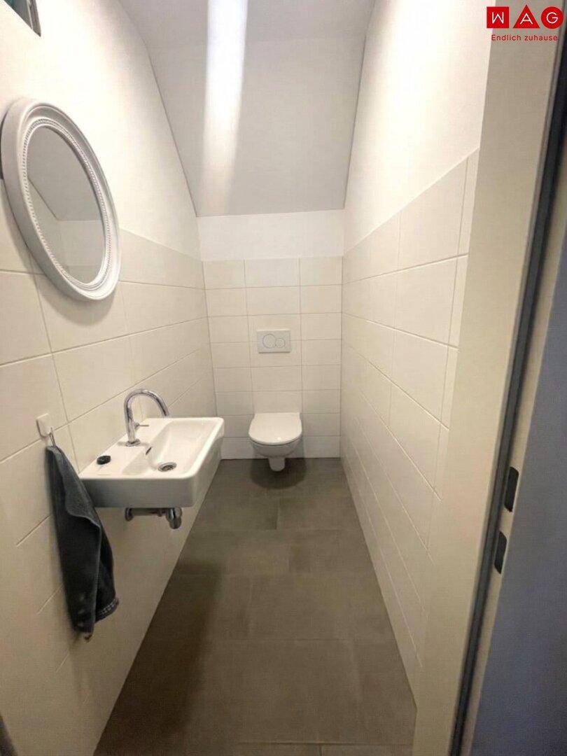 Kleines Gäste-WC mit wandhängender Toilette und Handwaschbecken, gefliest und mit Dachschräge.