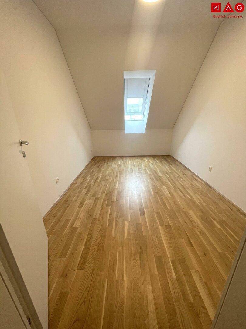 Leeres Zimmer mit Holzboden und einem Dachfenster, das den Raum unter der Schräge erhellt.