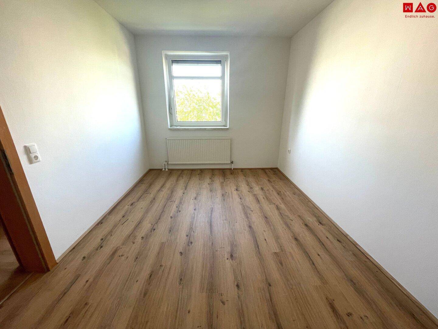 Leeres Schlafzimmer mit Laminatboden und einem Fenster, das viel Tageslicht hereinlässt.