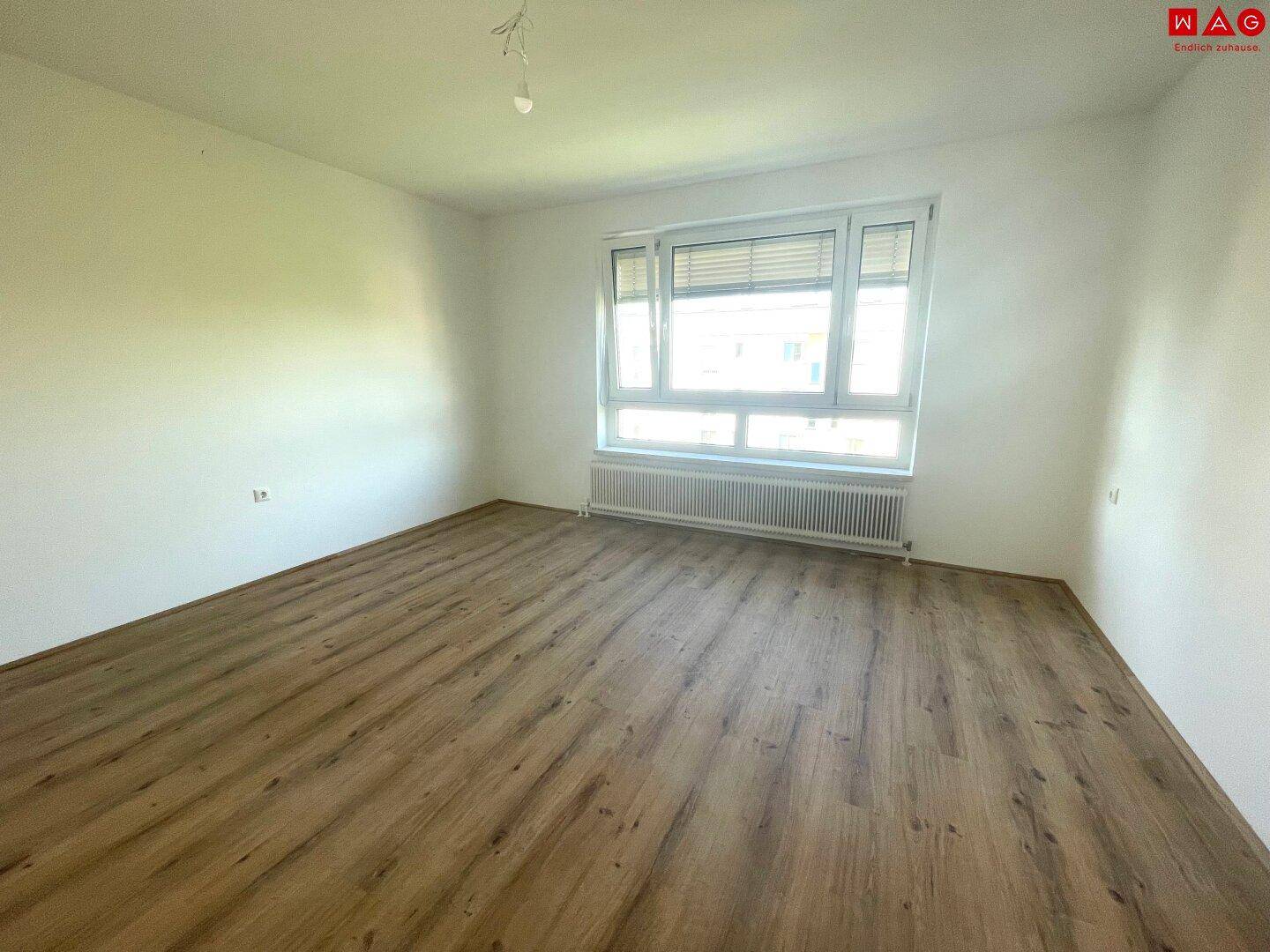 Geräumiges, leeres Wohnzimmer mit großem Fenster und Laminatboden, bereit zur Einrichtung.