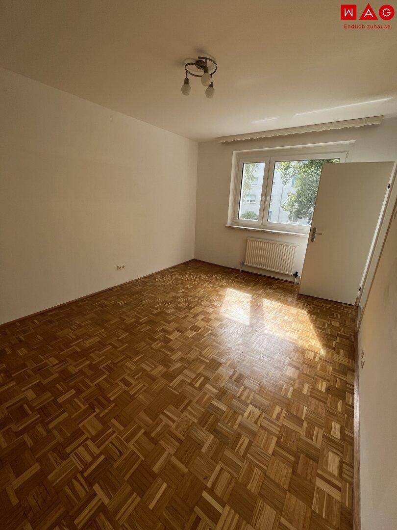 Leeres Schlafzimmer mit Parkettboden und Fenster, das viel Tageslicht hereinlässt.