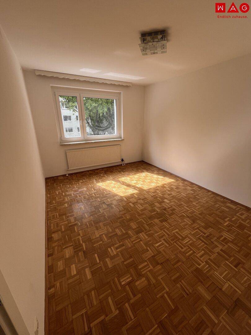 Unmöbliertes Schlafzimmer mit Parkettboden und einem Fenster, das Sonnenlicht hereinlässt.