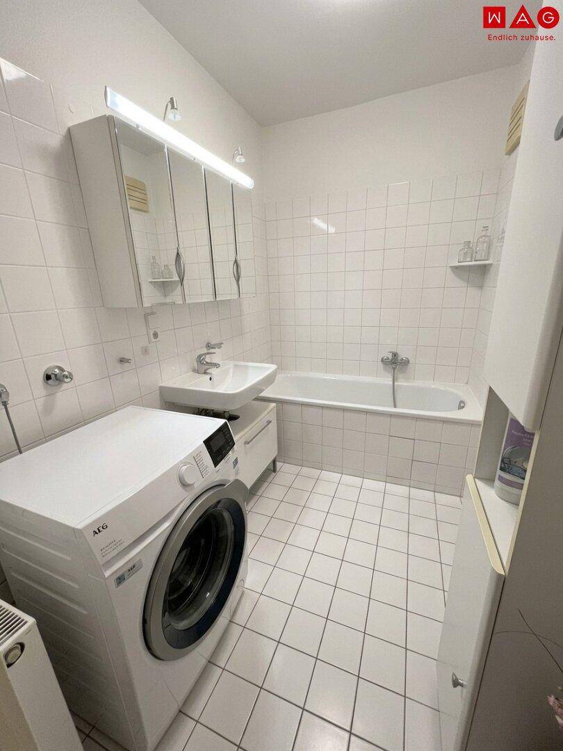 Funktionales Badezimmer mit Badewanne, Waschbecken und Waschmaschine, komplett gefliest in Weiß.
