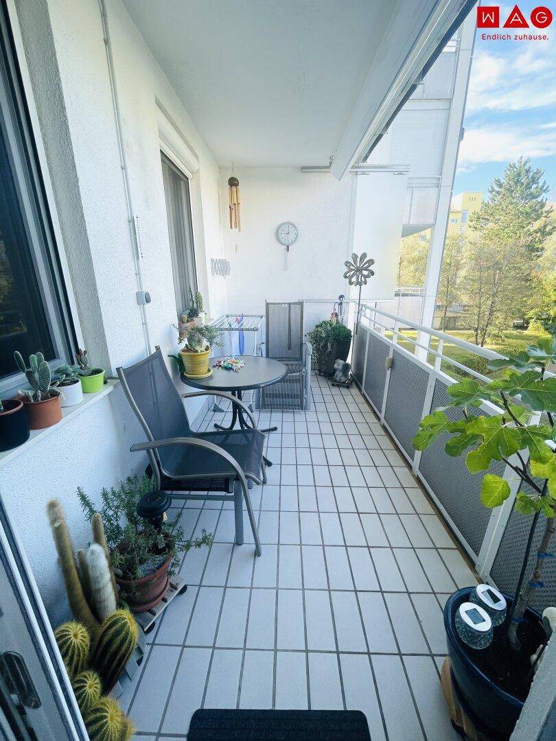 Sonnenverwöhnter Balkon mit Outdoor-Möbeln und Fliesenboden, umgeben von Pflanzen und Grünblick.