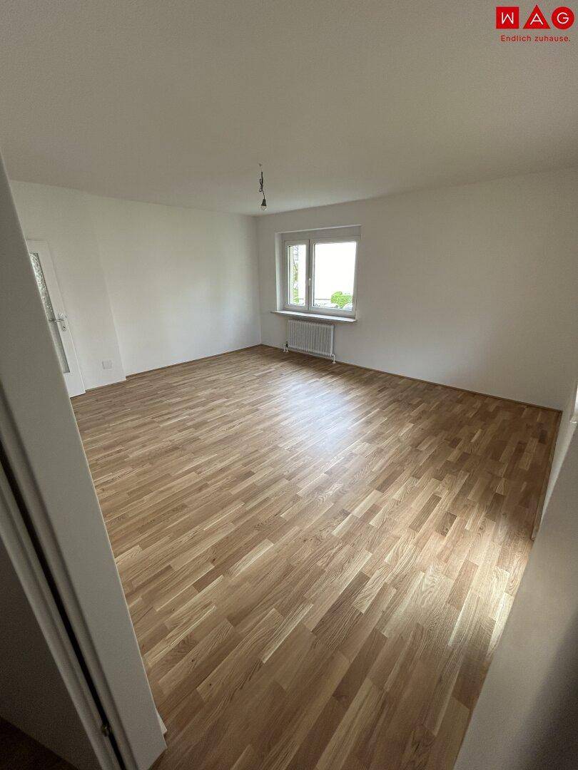 Geräumiges, leeres Zimmer mit hellem Parkettboden und großem Fenster, ideal als Schlafzimmer.