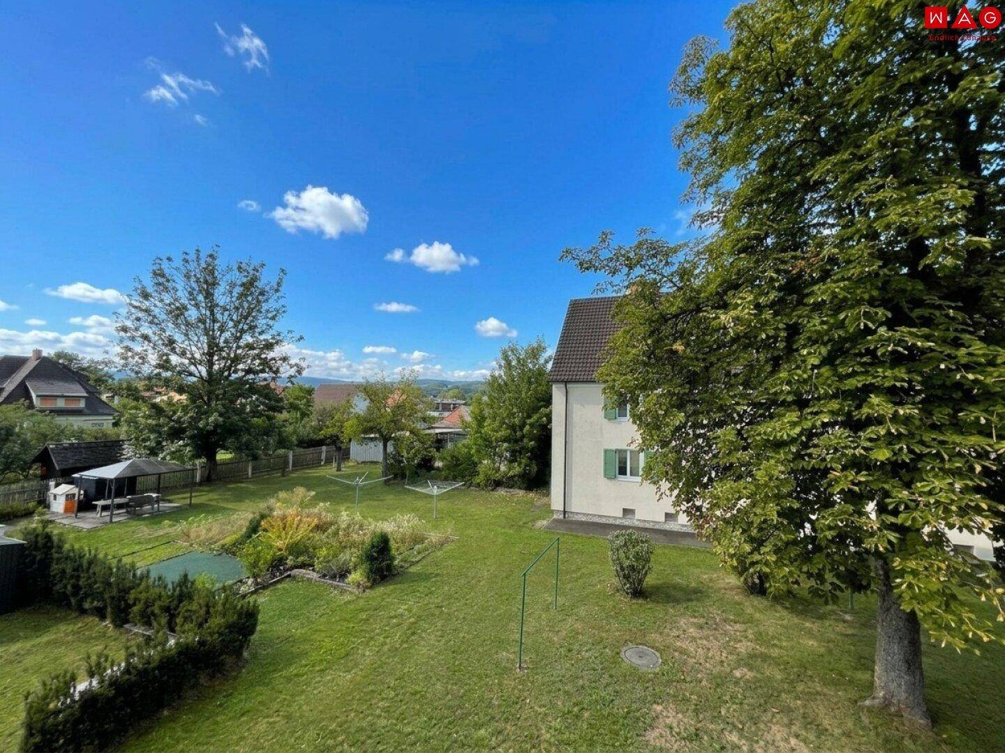 Großer Gemeinschaftsgarten mit Rasenfläche, Bäumen und einem Pavillon unter strahlend blauem Himmel.