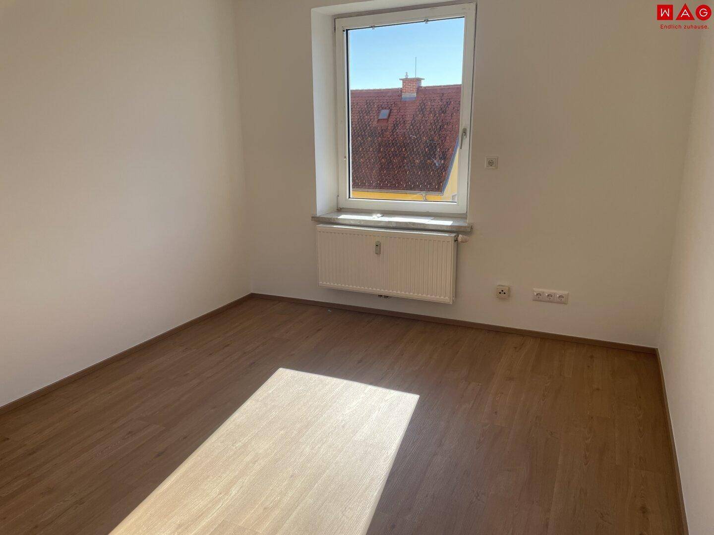 Helles, leeres Zimmer mit Laminatboden und Fenster, das viel Tageslicht hereinlässt.