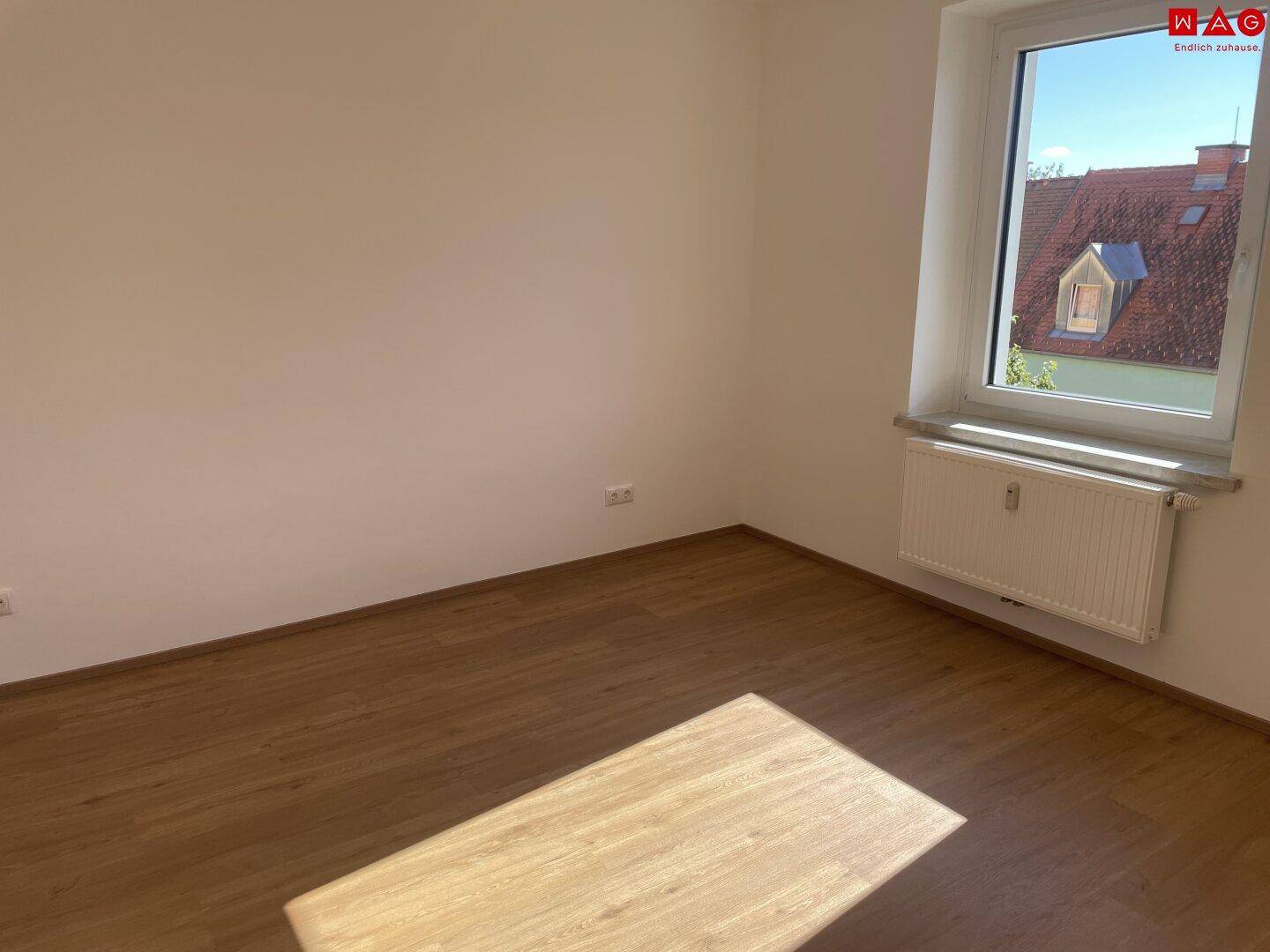 Lichtdurchflutetes Zimmer mit Laminatboden und Ausblick auf die umliegenden Dächer.