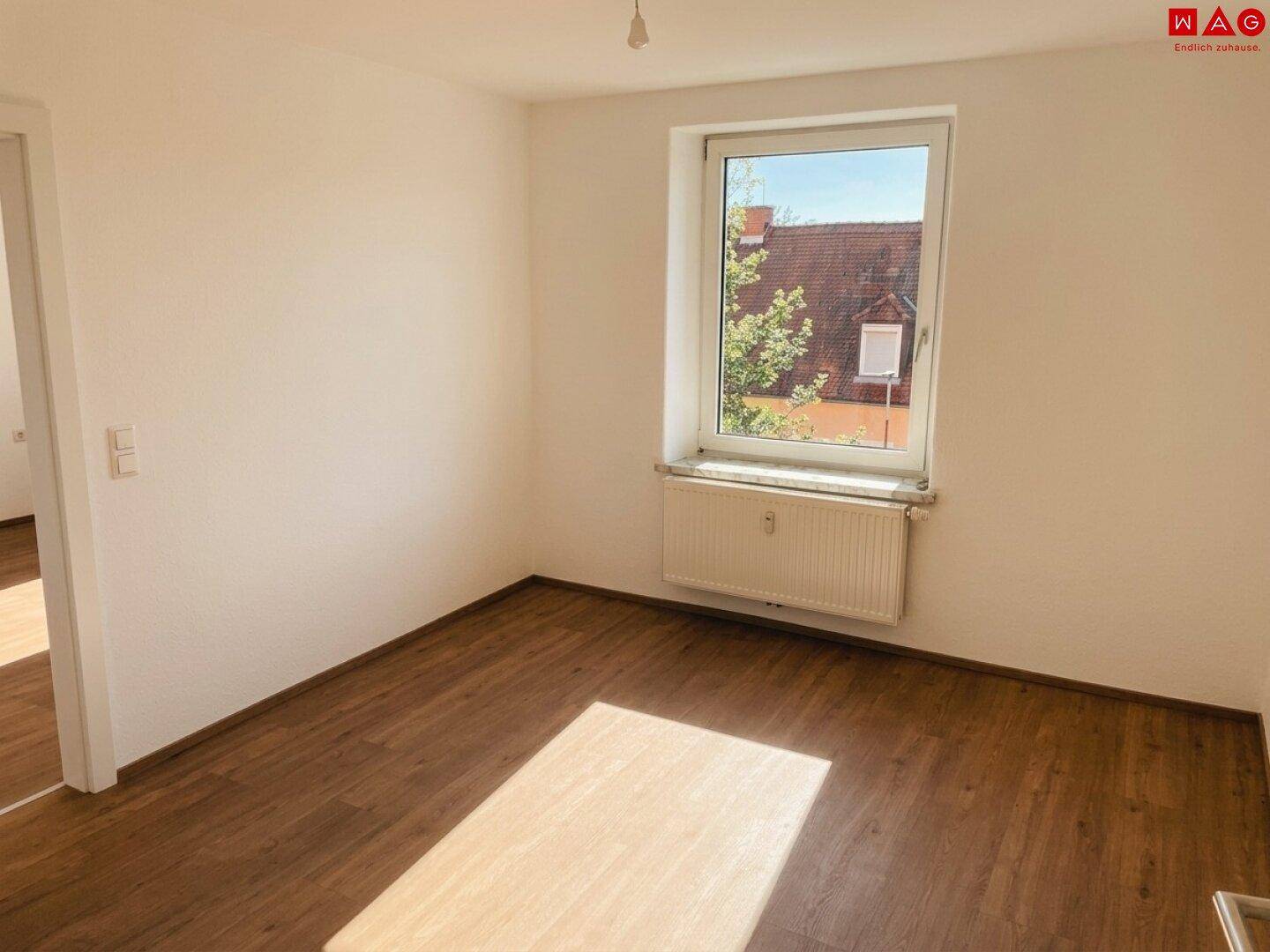 Leeres Zimmer mit Holzboden, großem Fenster und Blick auf die umliegenden Dächer.