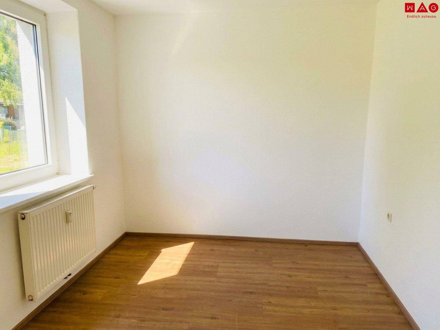 Helles, leeres Zimmer mit Holzboden und Fenster mit Blick ins Grüne.