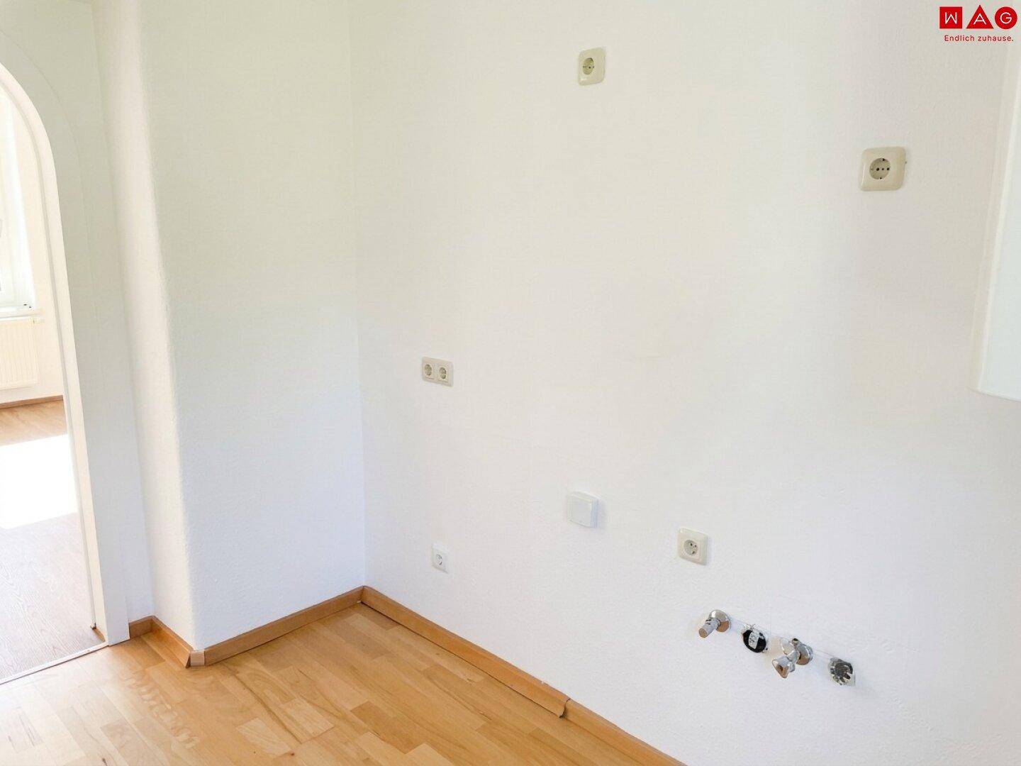 Leerer Raum mit Holzboden und Anschlüssen für eine Küche, verbunden durch einen Bogen.