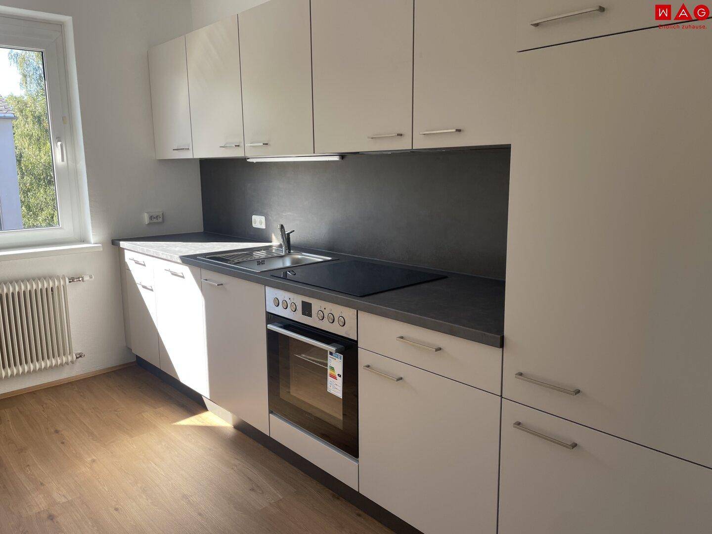 Helle Einbauküche in Grau mit dunkler Arbeitsplatte, integrierten Geräten und einem Fenster für Tageslicht.