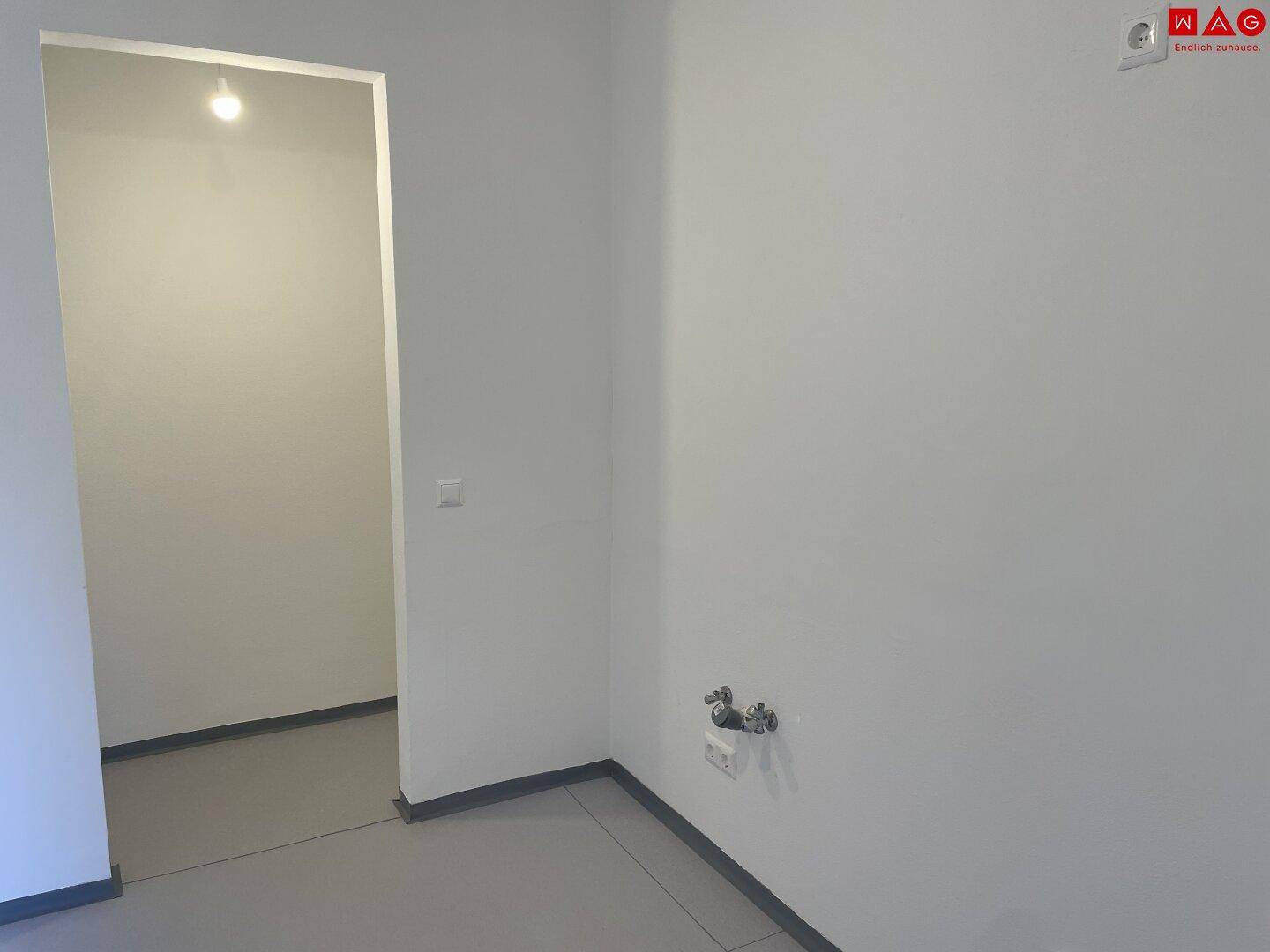 Leerer Raum mit Fliesenboden und Anschlüssen für eine zukünftige Küche oder Waschmaschine.
