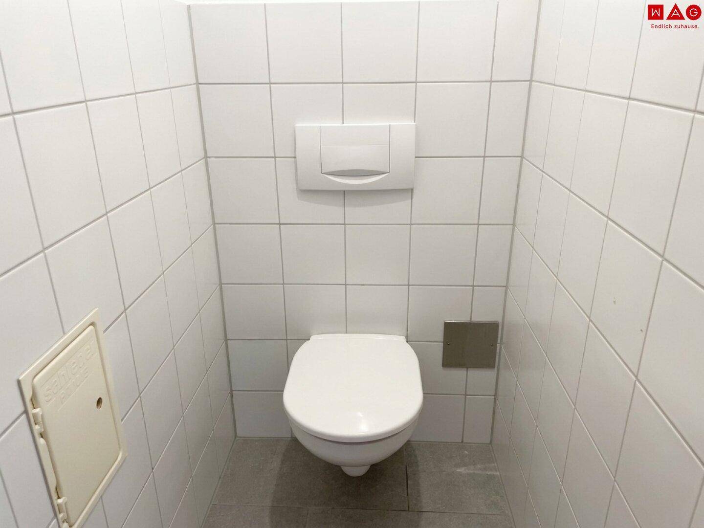 Separates WC mit wandhängender Toilette und weißen Fliesen an den Wänden.