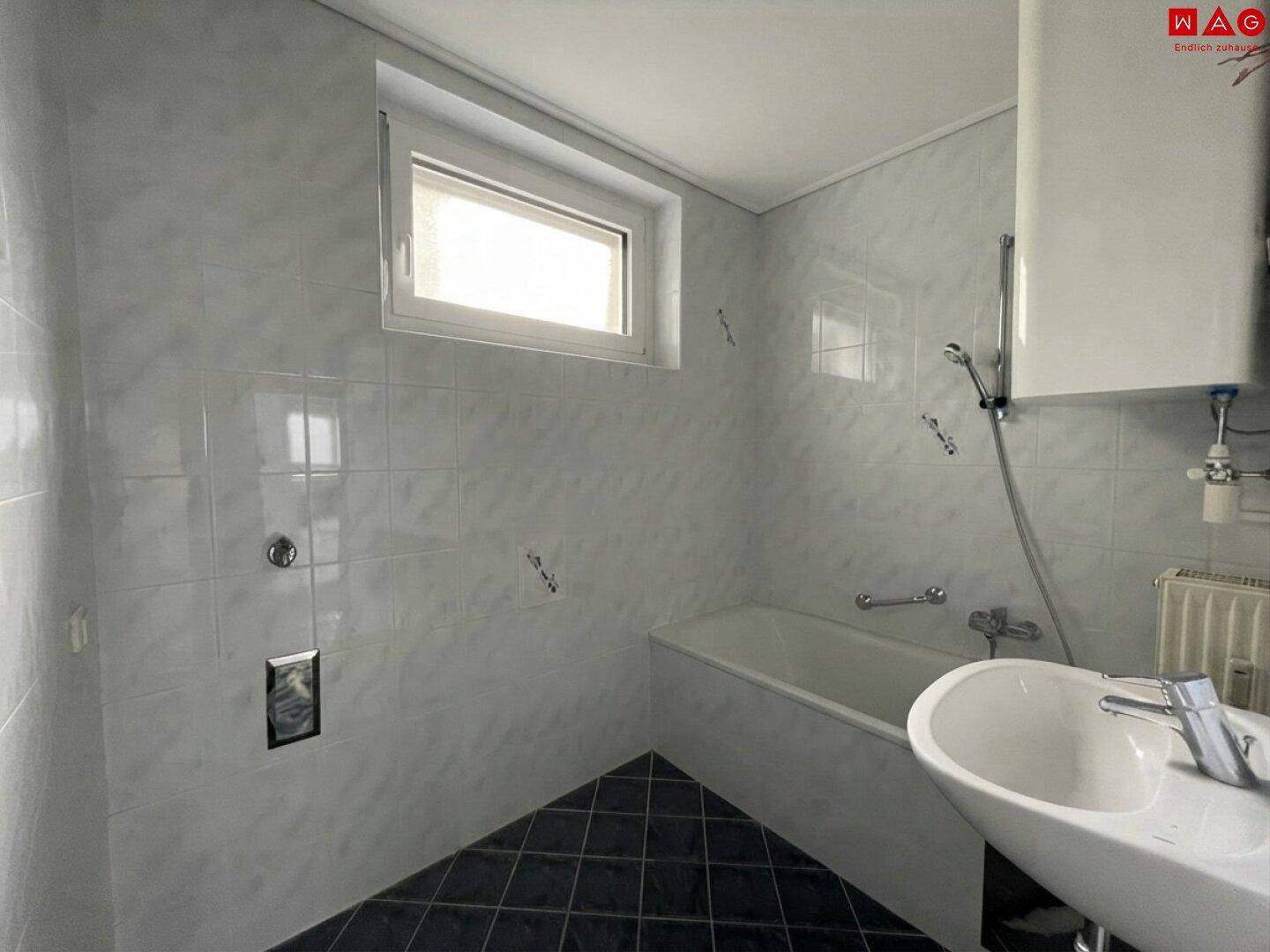 Badezimmer mit Badewanne, Waschbecken, Fenster und weißen Fliesen an den Wänden.