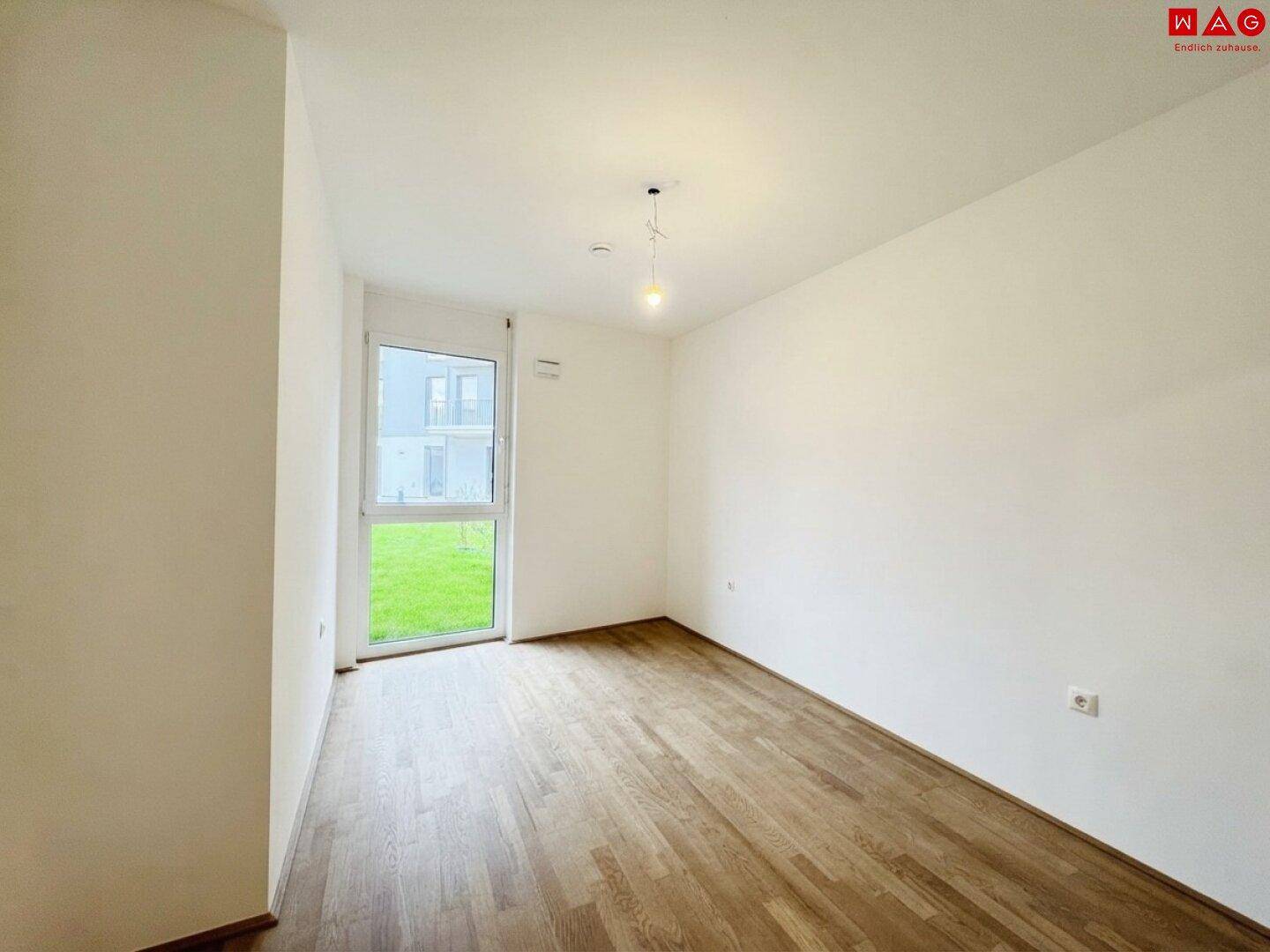 Freundliches Zimmer mit Holzboden und großer Fenstertür, die viel Tageslicht hereinlässt.