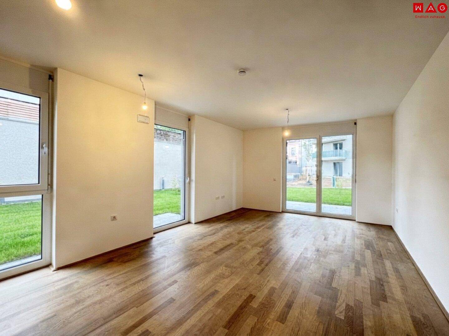 Heller Wohnraum mit Holzboden und großen Fenstertüren, die Zugang zur Grünfläche bieten.