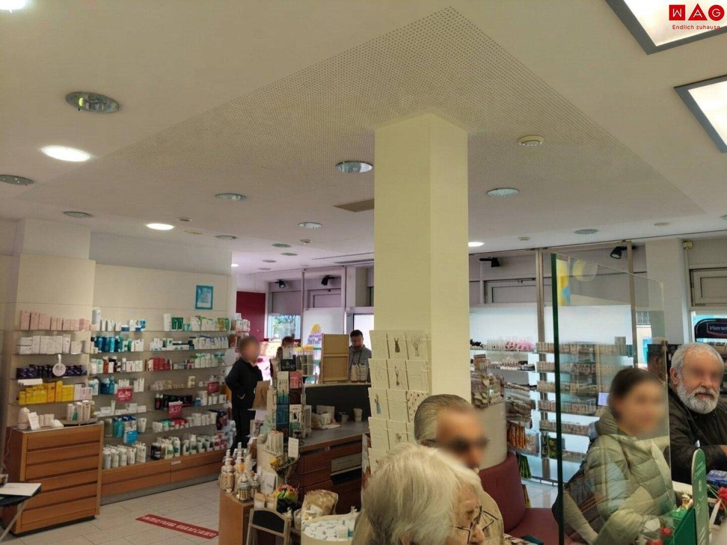 Innenansicht einer Apotheke mit Kunden, Produktregalen und einer Säule in der Mitte des Raumes.