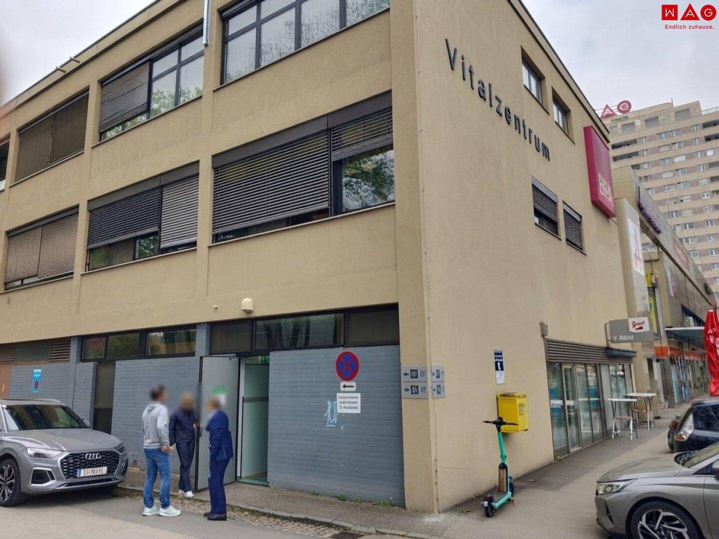 Seitenansicht des Vitalzentrums mit Parkplätzen und Eingangsbereich, umgeben von städtischer Infrastruktur.