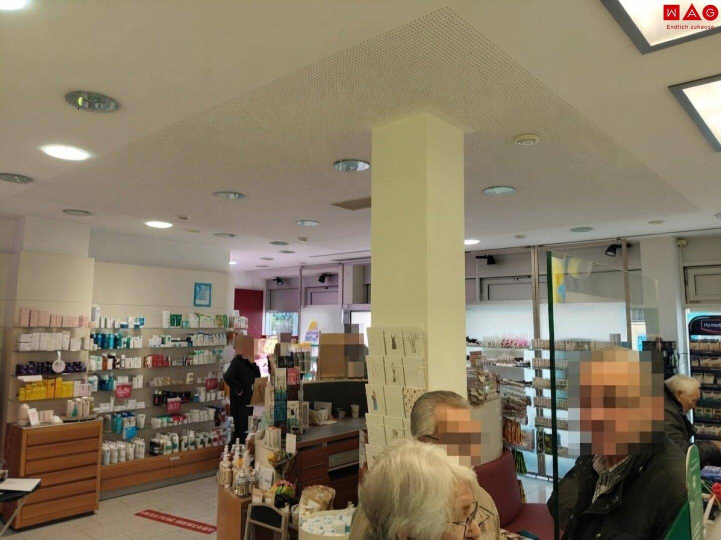 Verkaufsraum einer Apotheke mit Säule, Deckenbeleuchtung und Kunden im Hintergrund.