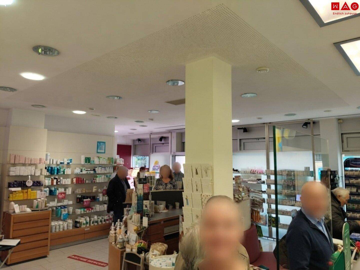 Innenansicht einer Apotheke mit Verkaufsregalen, einem zentralen Pfeiler und Kunden im Laden.