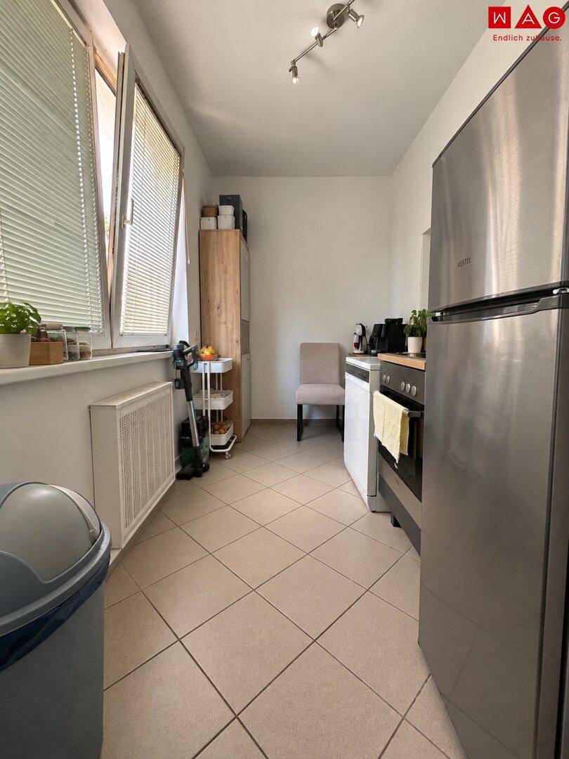 Schmale Küche mit Fliesenboden, Fenster und grundlegenden Küchengeräten wie Kühlschrank und Ofen.