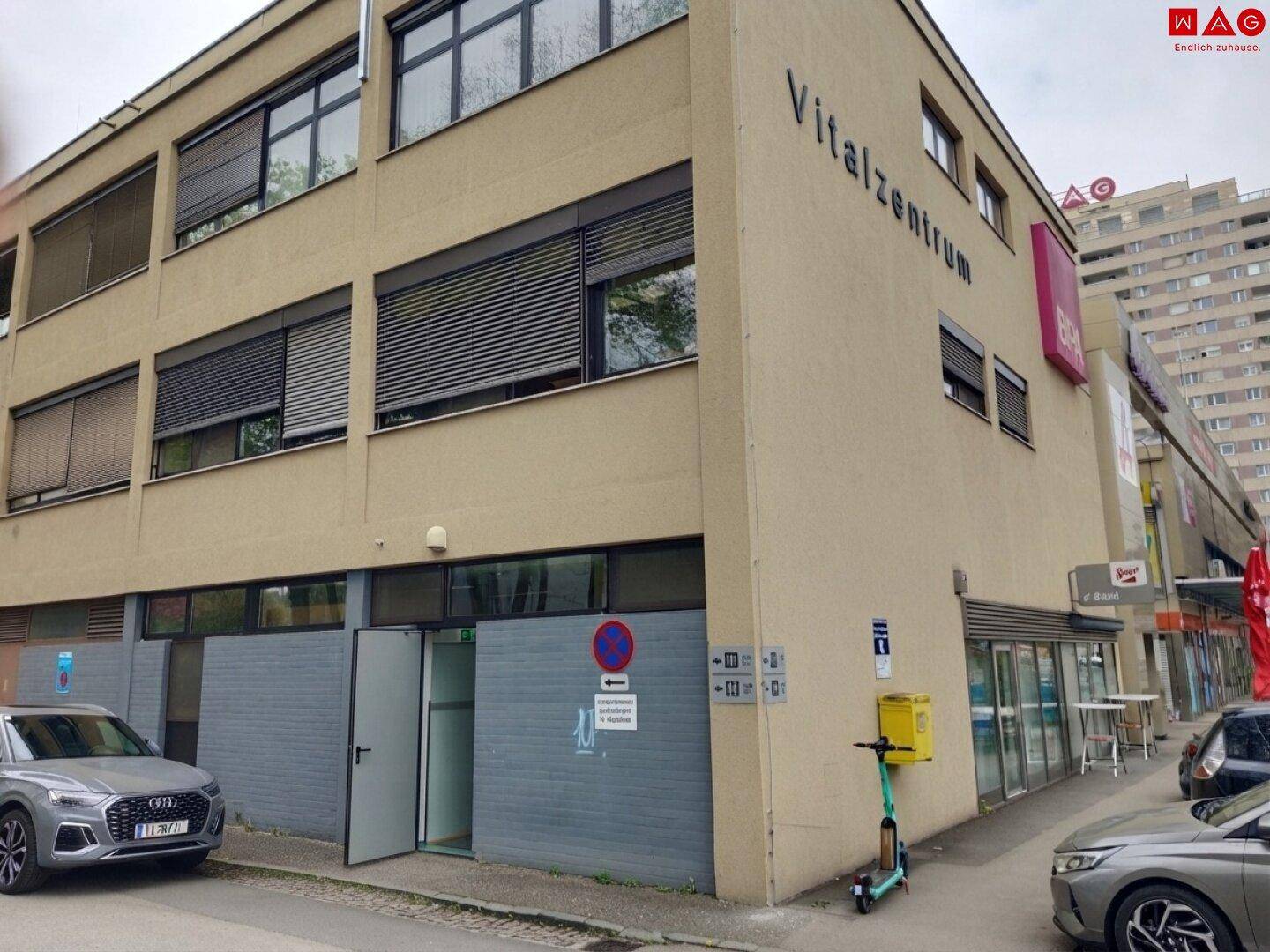 Seitenansicht des Vitalzentrums mit Geschäften im Erdgeschoss und geparkten Autos auf der Straße.