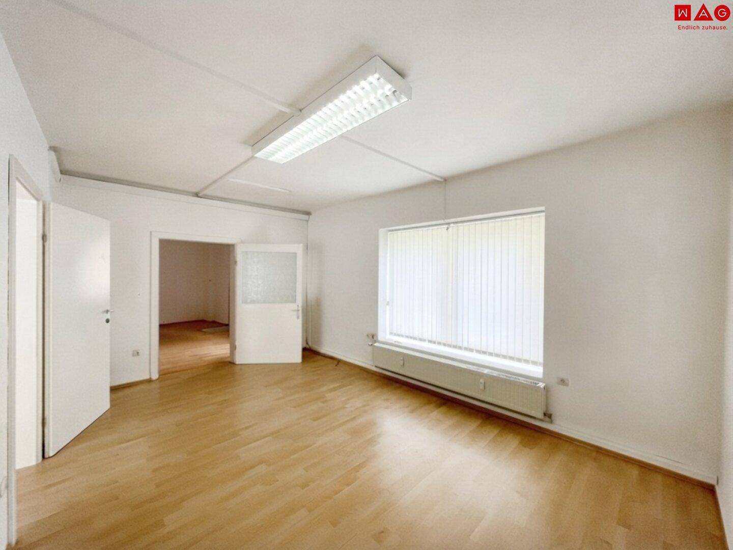 Heller, leerer Raum mit hellem Holzparkettboden, weißen Wänden und einem großen Fenster mit Jalousien.