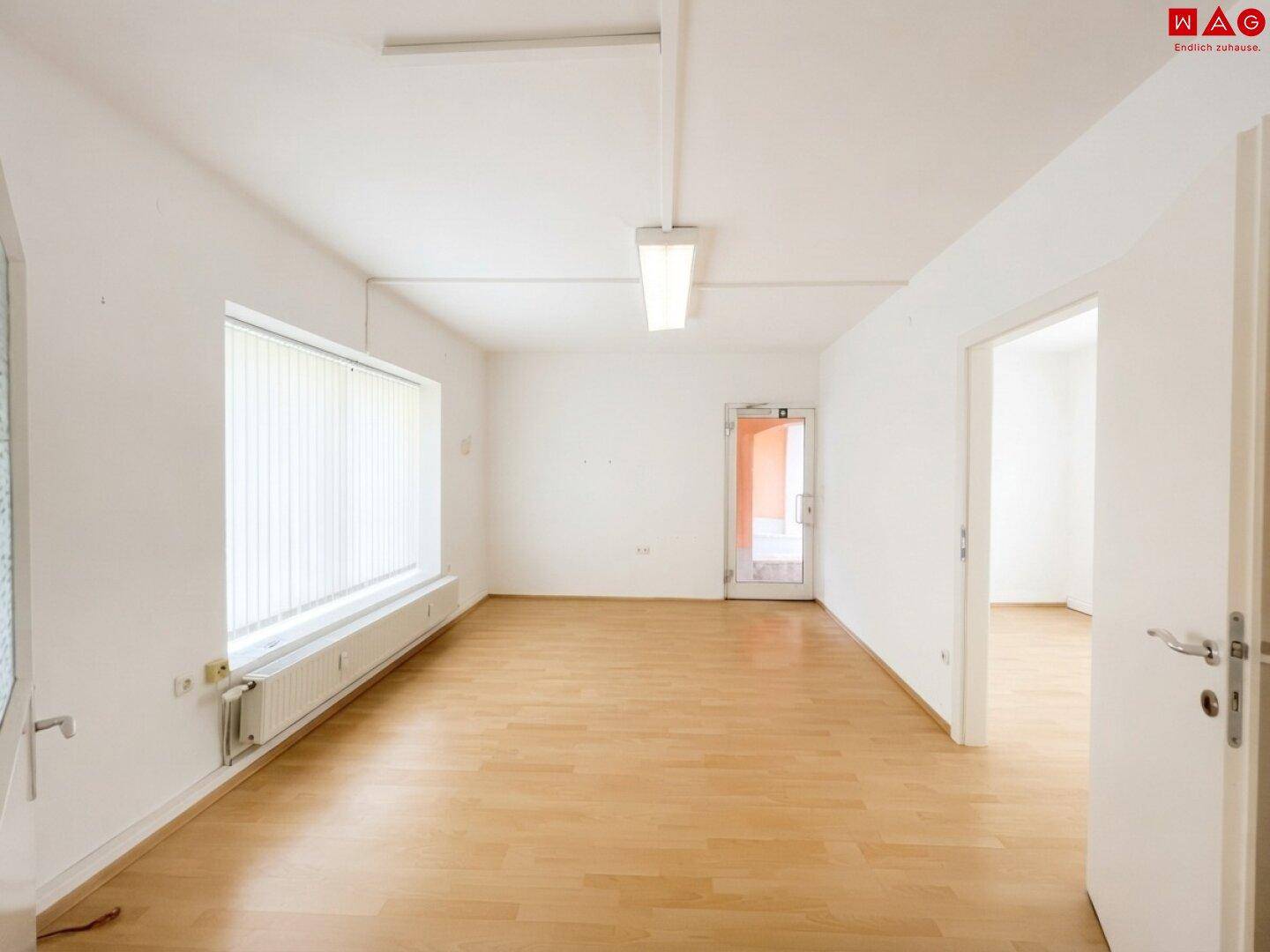 Geräumiger, leerer Raum mit hellem Holzboden, weißen Wänden und einem Fenster mit vertikalen Jalousien.