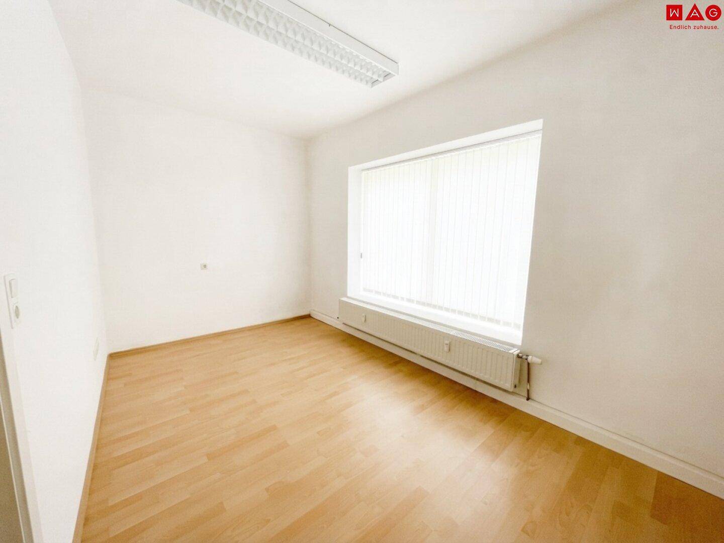 Kleinerer, leerer Raum mit Holzboden, weißen Wänden und einem Fenster mit Jalousien für natürliches Licht.