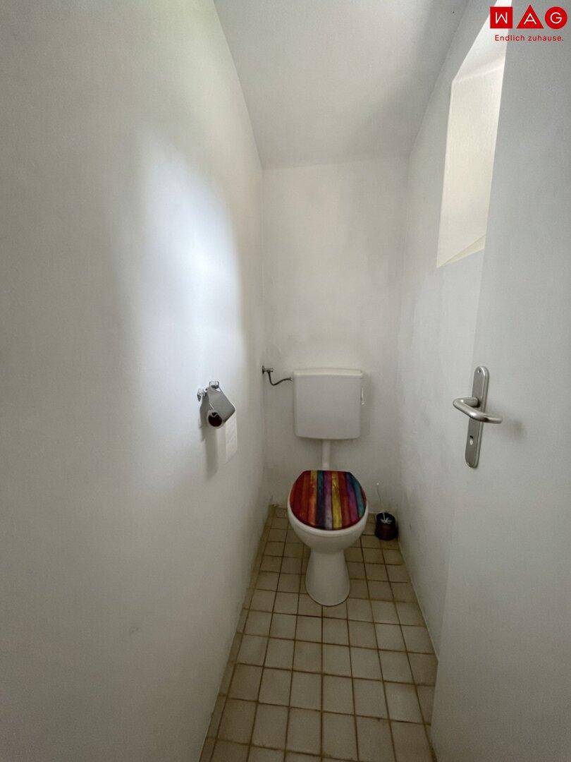 Schmale Toilette mit Fliesenboden, weißer Keramik und einem kleinen Fenster für Belüftung.