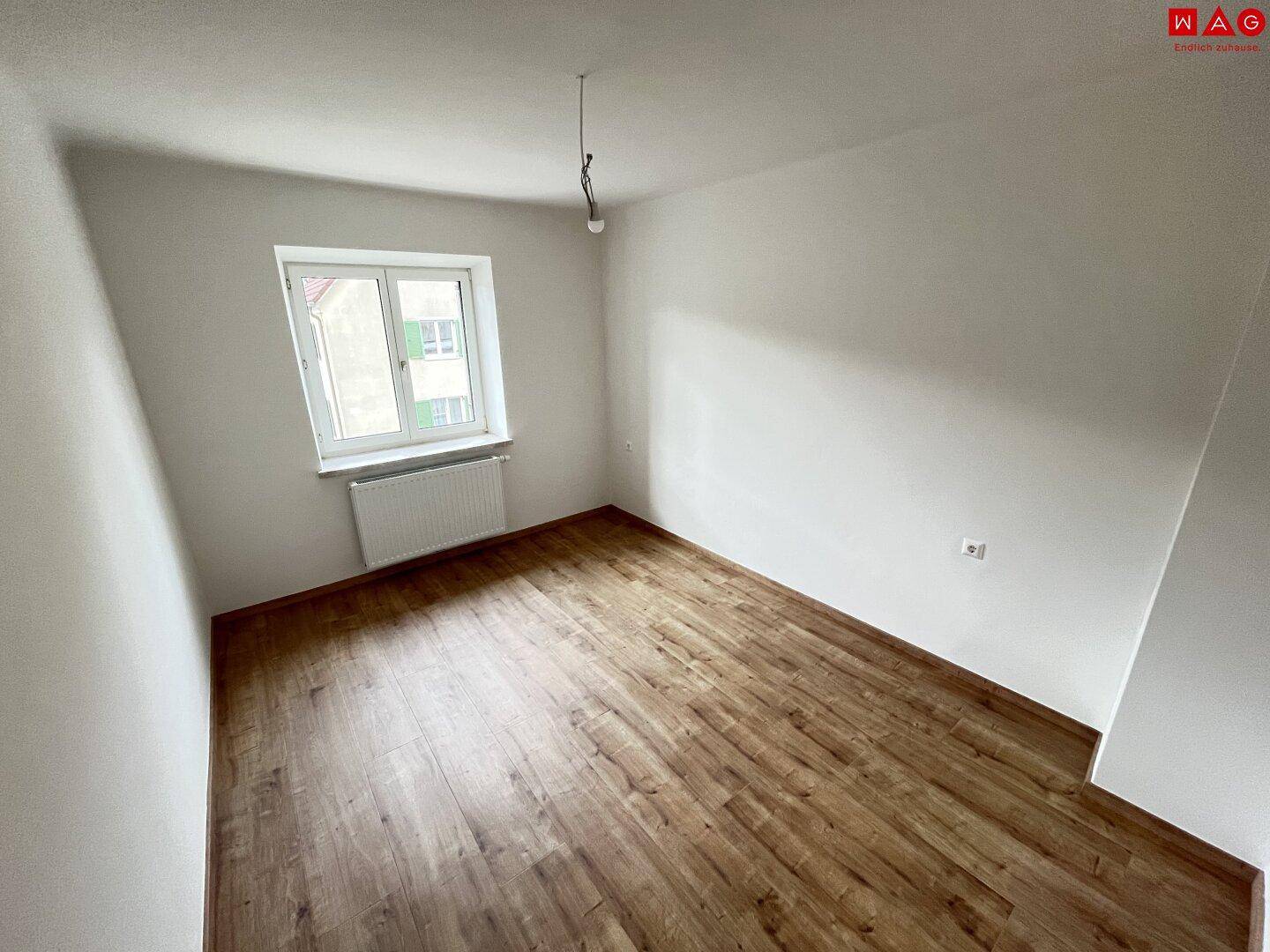 Leeres Zimmer mit hellem Laminatboden, weiß gestrichenen Wänden und einem Heizkörper unter dem Fenster.