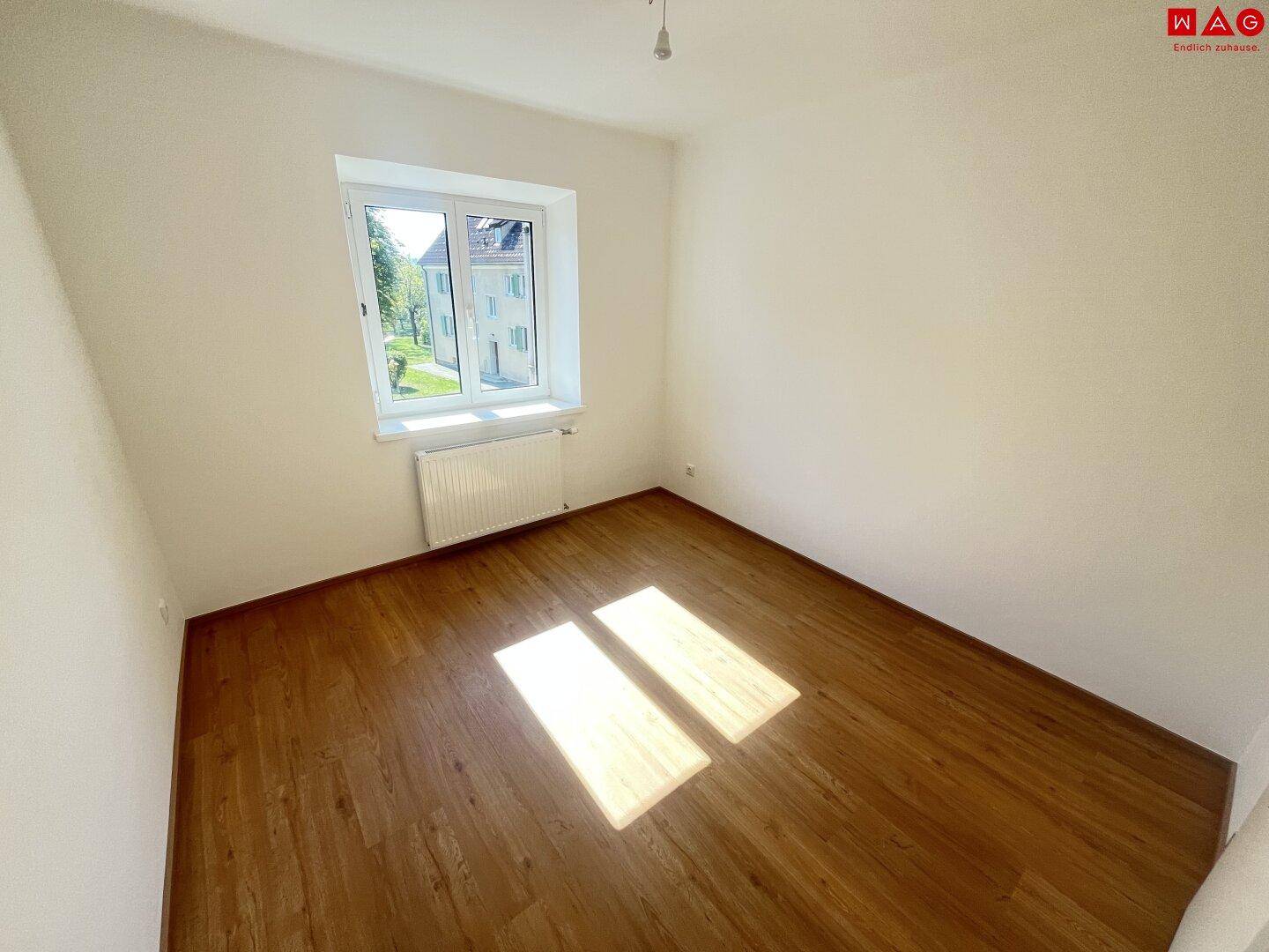 Sonniger Raum mit warmem Parkettboden und großen Fenstern, die einen Blick ins Grüne bieten.