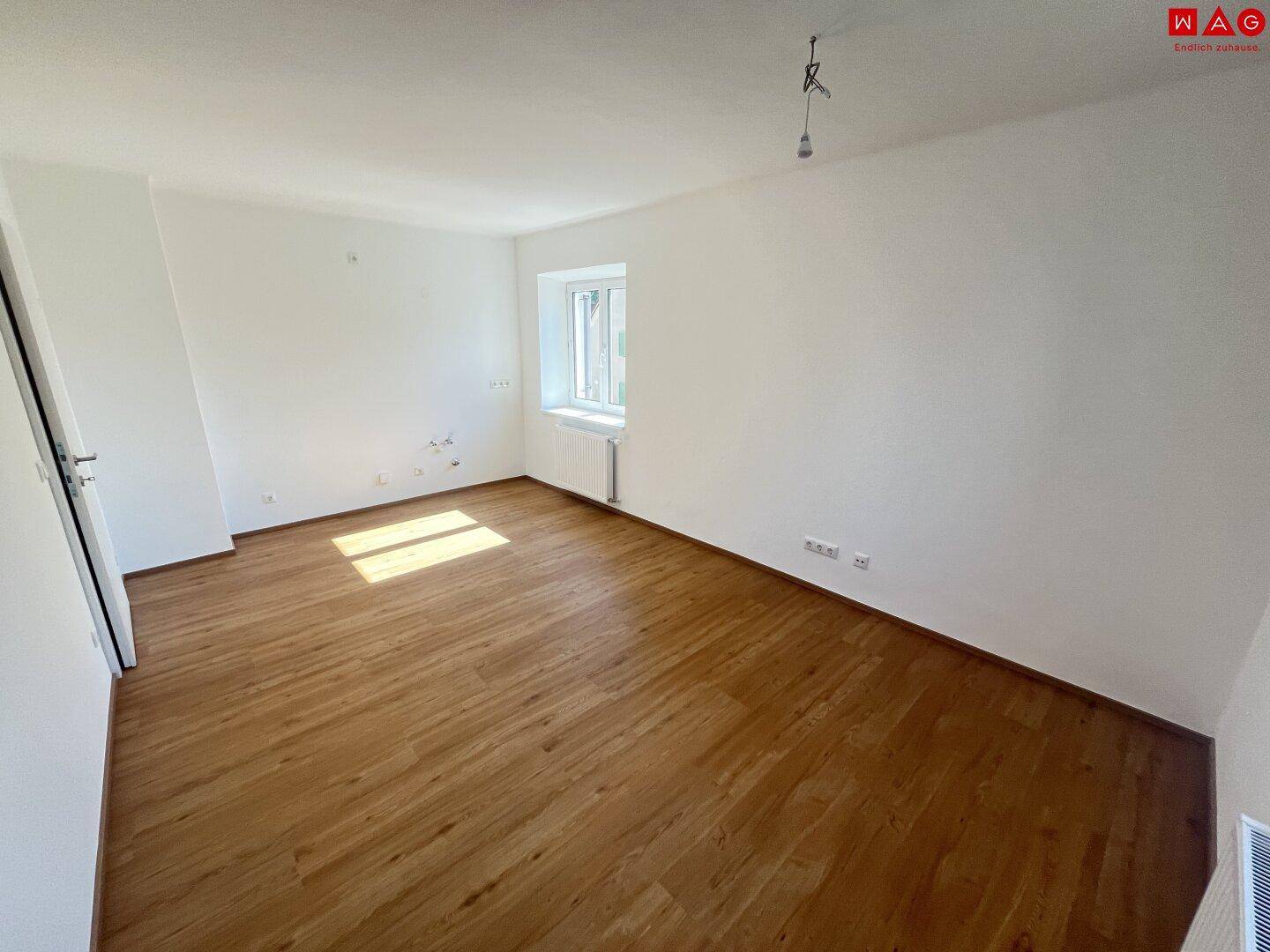 Heller, leerer Raum mit Parkettboden, weißen Wänden und einem Fenster, das Sonnenlicht hereinlässt.