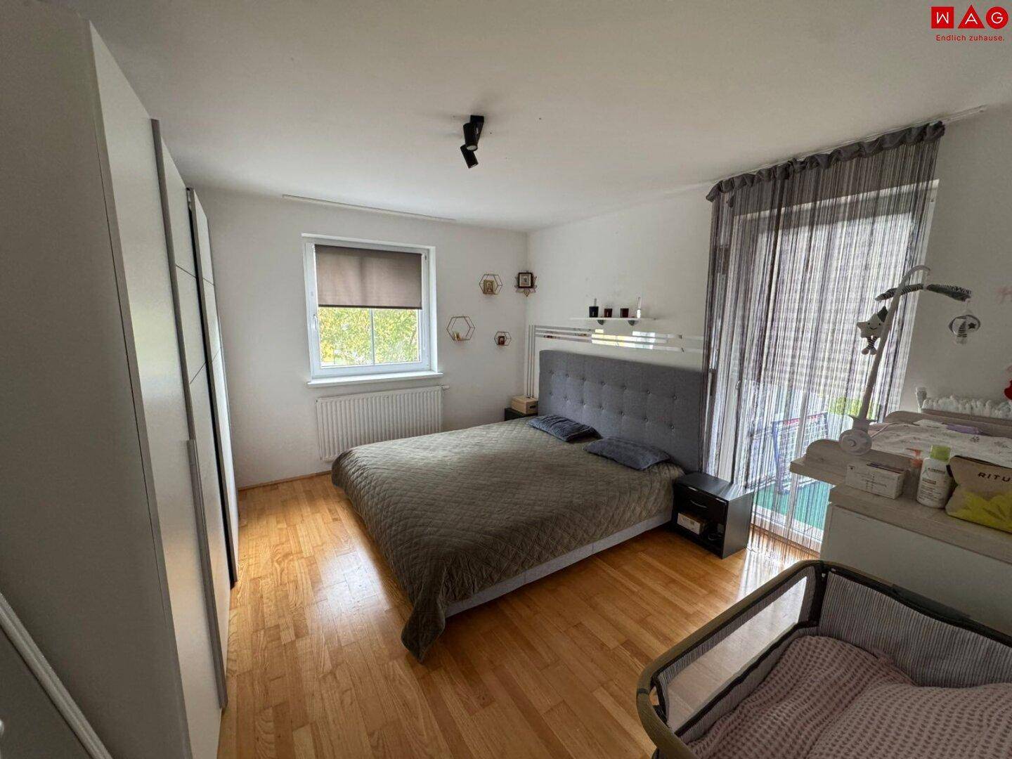 Schlafzimmer mit Doppelbett, Kleiderschrank und Babybett, ausgestattet mit Holzboden und Balkonzugang.