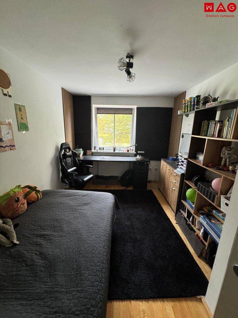 Jugendzimmer mit Bett, Schreibtisch und Regal, ausgestattet mit Holzboden und einem dunklen Teppich.