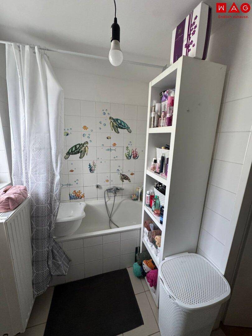 Badezimmer mit Badewanne und Duschvorhang, weißen Fliesen und einem Fenster für Tageslicht.