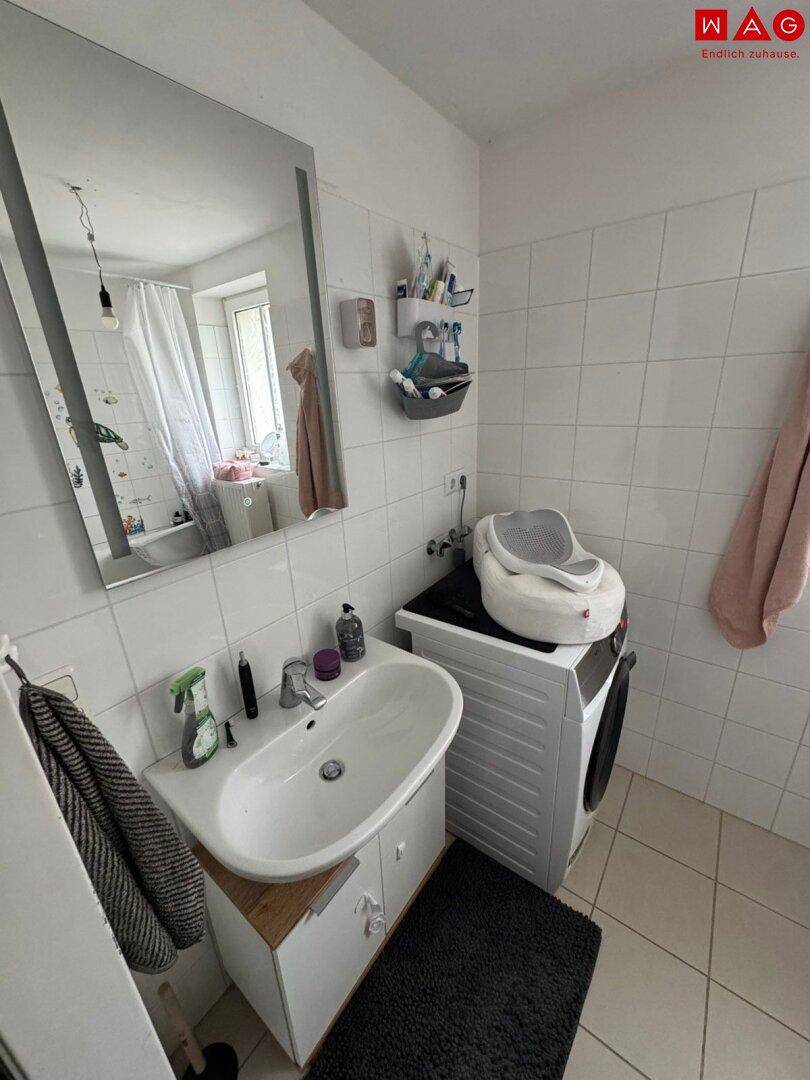 Badezimmer mit Waschbecken, Spiegel und Waschmaschine, ausgestattet mit weißen Fliesen und Tageslicht.