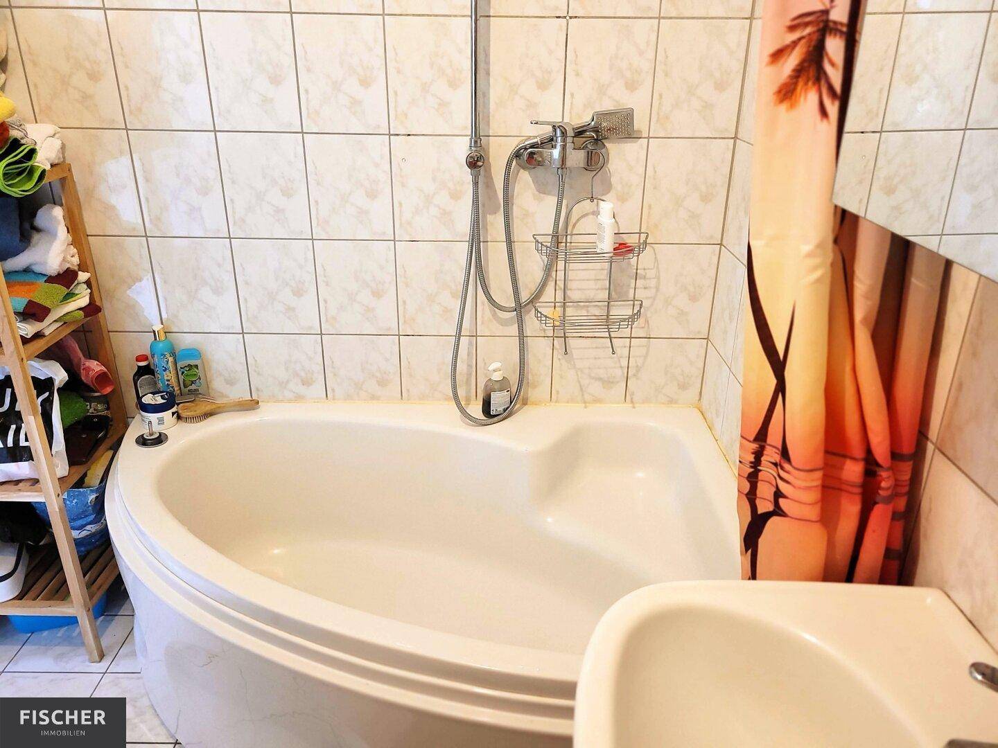 Badezimmer mit Eckbadewanne und Duschvorrichtung, gefliesten Wänden und kleinem Waschbecken.