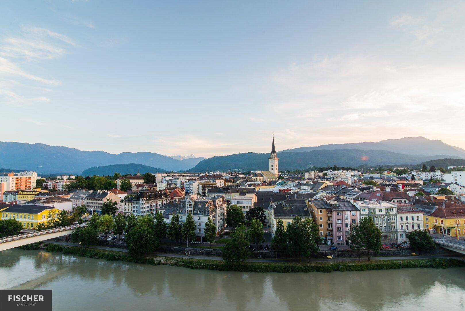 Panoramablick auf eine Stadt am Fluss mit Bergen im Hintergrund unter einem klaren blauen Himmel.