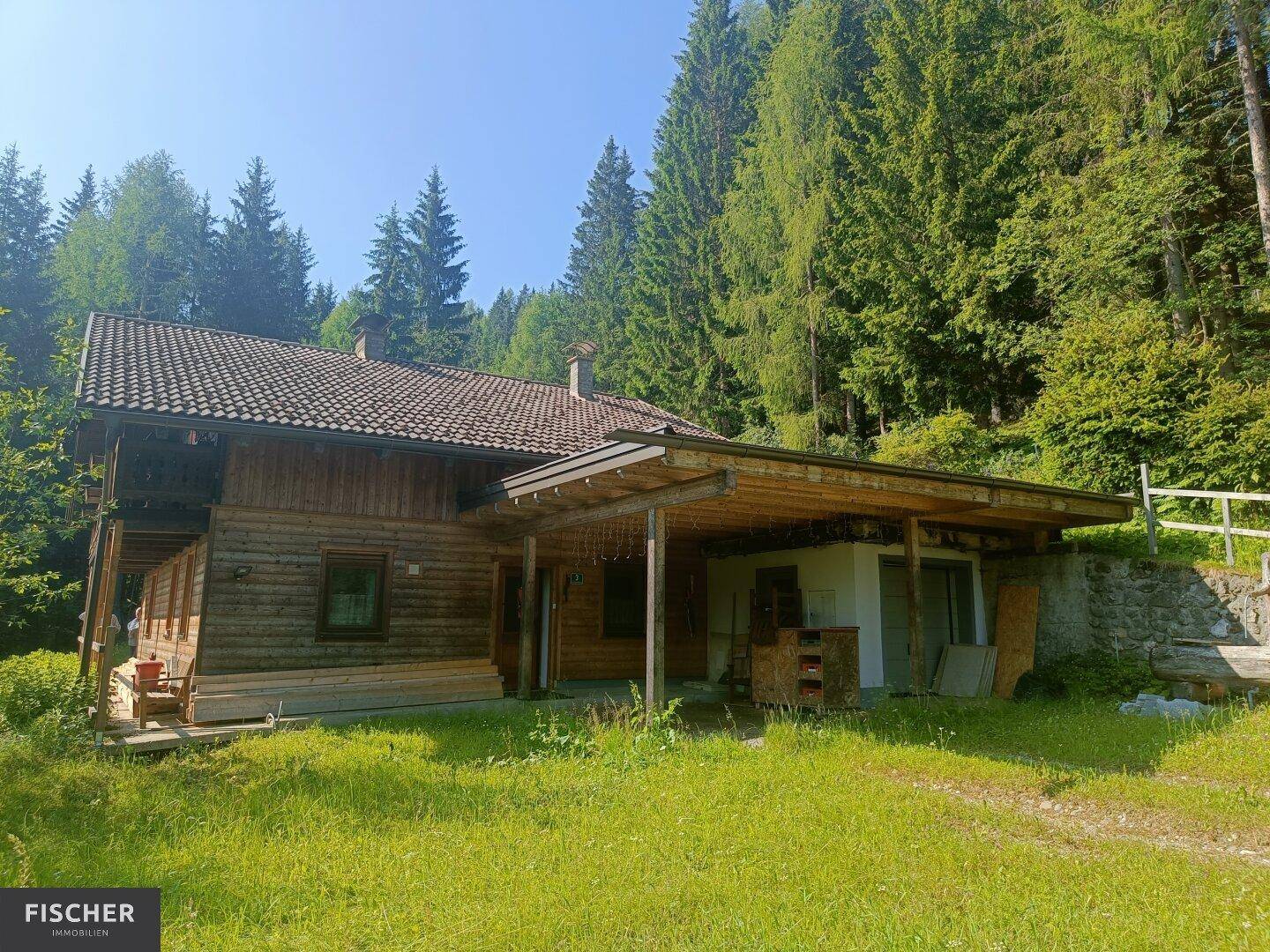 Rustikales Holzhaus mit Carport, umgeben von Wald und Wiesen in sonniger alpiner Lage.
