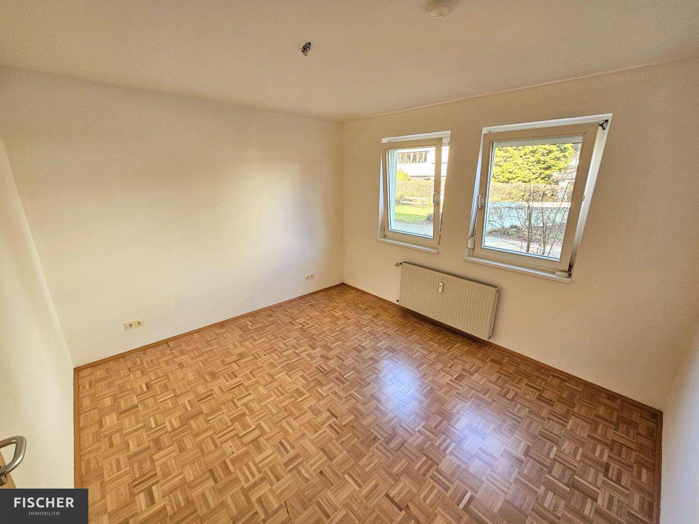 Helles Schlafzimmer mit Parkettboden und zwei Fenstern, die viel natürliches Licht hereinlassen.