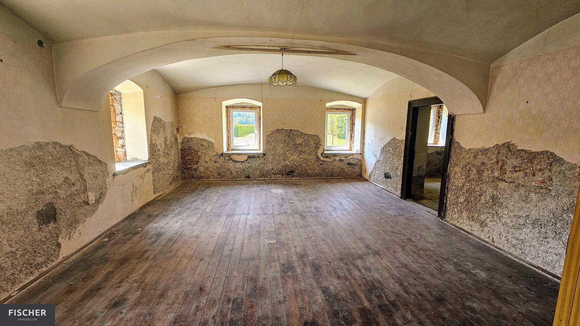 Großer Raum mit gewölbter Decke, abblätternder Tapete und altem Holzboden, renovierungsbedürftig.