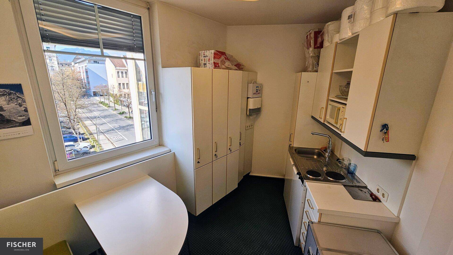 Kleine Teeküche mit Spüle, Kochplatten, weißen Schränken und Blick auf die Straße durch ein Fenster.