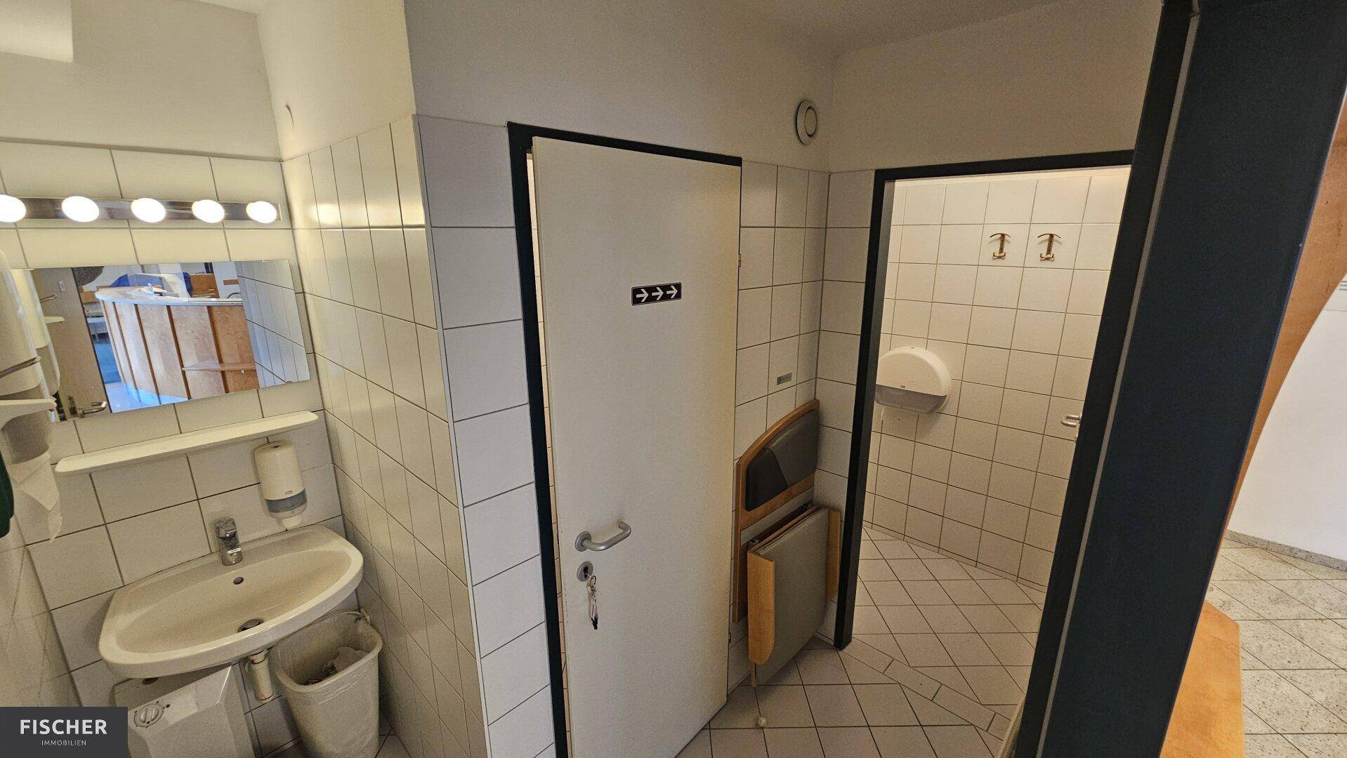Sanitärbereich mit Waschbecken, Spiegel und gefliesten Wänden, sowie Zugang zu weiteren Toiletten.