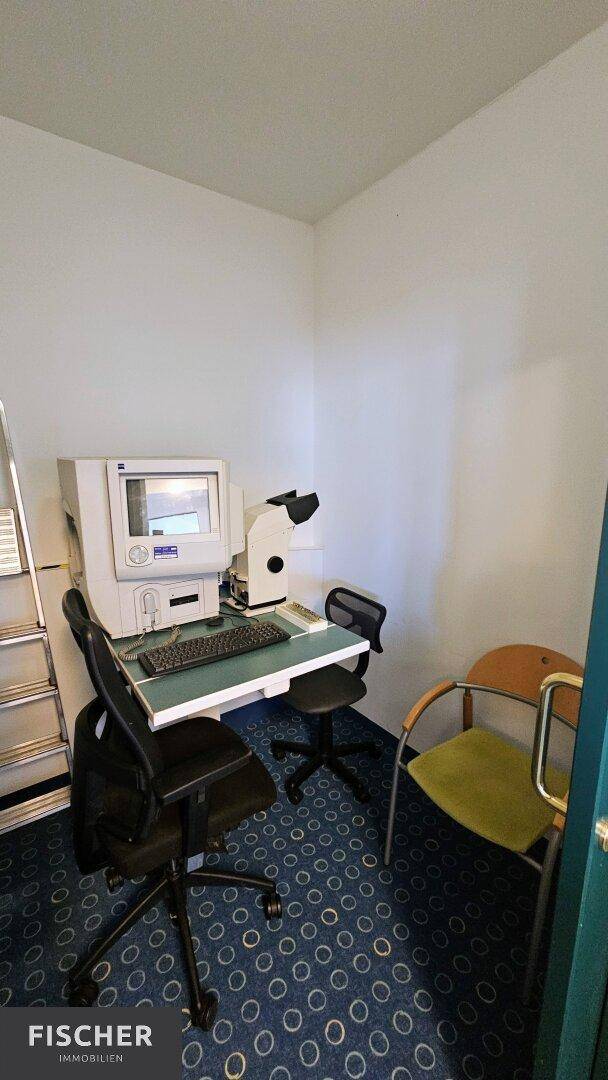 Kleiner Arbeitsraum mit medizinischem Gerät, Schreibtisch und Bürostuhl, ausgestattet mit Teppichboden.