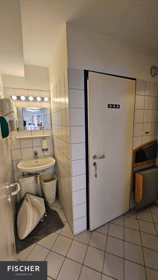 Toilettenraum mit weiß gefliesten Wänden, Waschbecken und Spiegel mit Beleuchtung in einer Praxis.