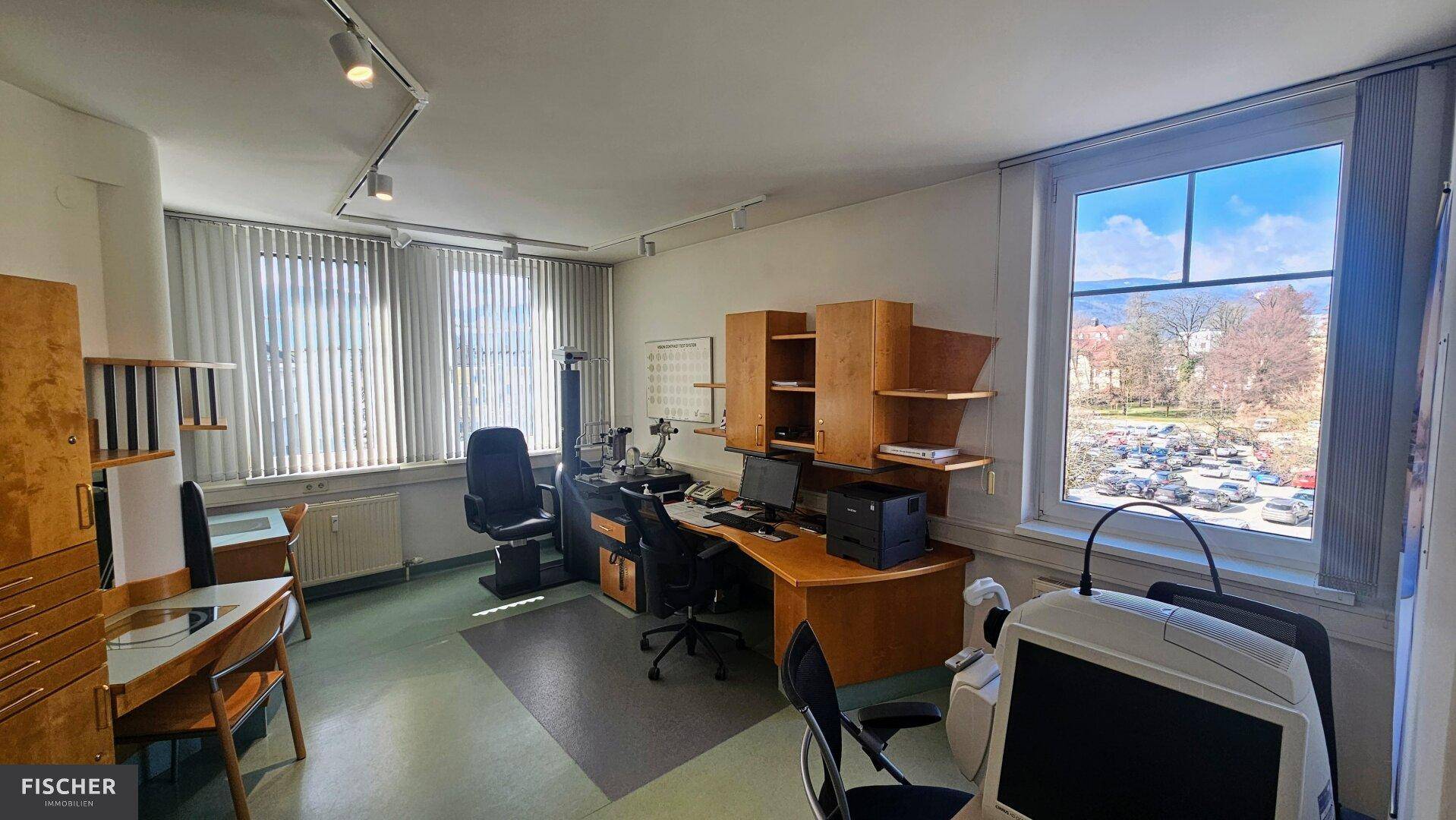 Behandlungszimmer mit Schreibtisch, Bürostühlen und großem Fenster mit Jalousien und Blick ins Grüne.